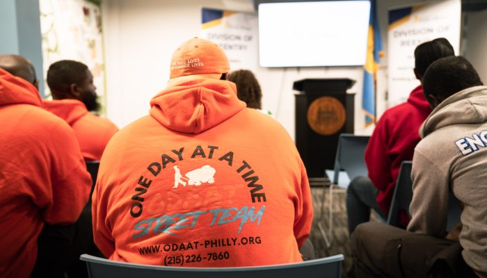 Individuals sit in front of a podium to listen to an event about justice-impacted people and voting. A man's sweatshirt says 'One Day at a Time'