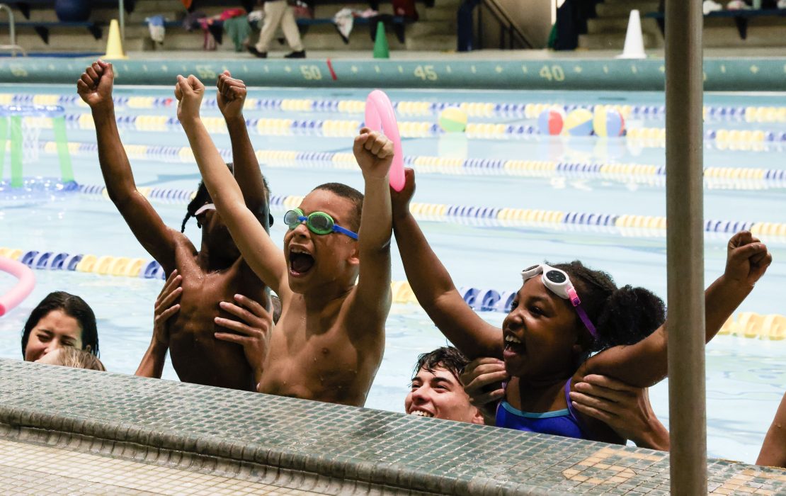 Children are gathered at the side of a pool cheering.