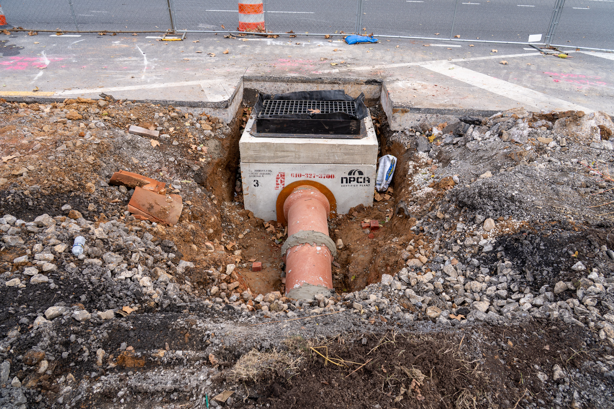 Uno de los nuevos desagües pluviales fotografiado durante la construcción con el suelo que lo rodea excavado. El desagüe presenta una rejilla metálica pintada de negro en la parte superior de una gran caja de hormigón colocada en el suelo. Una tubería grande de color rojo pálido se conecta al costado de la caja de concreto y continúa bajo tierra.