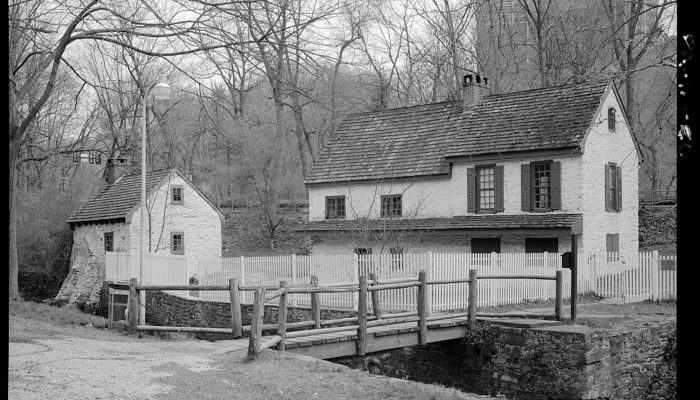 Black and white photo of two white-washed stone buildings with footbridge in the foreground. A tall, modern, apartment building can be seen from a distance through the trees.