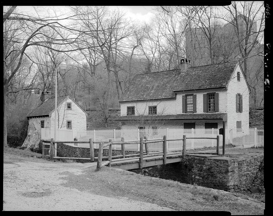 Black and white photo of two white-washed stone buildings with footbridge in the foreground. A tall, modern, apartment building can be seen from a distance through the trees.