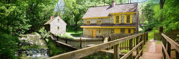 Color photo of two stone buildings with bright yellow trim. A footbridge appears across a small stream in the foreground.
