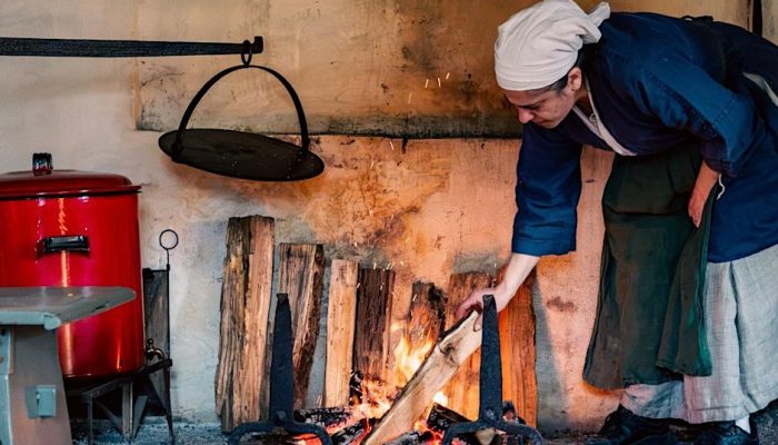 A color photo of a museum educator building a fire in a historic hearth.