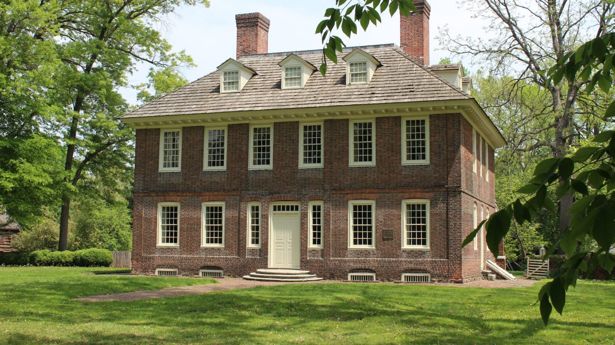 A two-and-a-half story brick building with light trim and wood-shake roof. Purple flowers appear in the foreground.