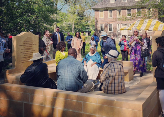 Color photo of people sitting on bench components of a memorial. Standing people, a yellow event tent, and a brick building are visible in the background.