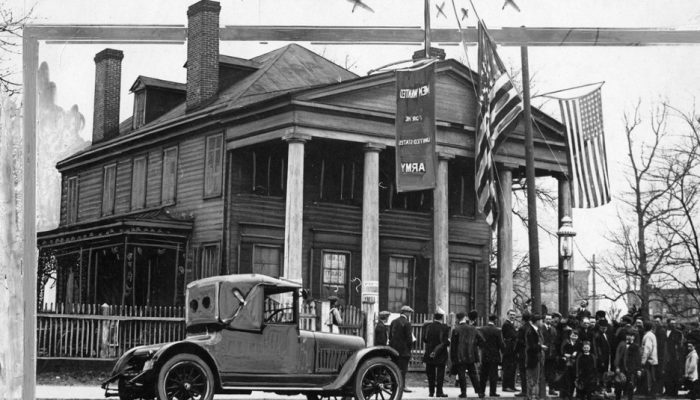 A black and white photo of a two-story house with four columns in the front facade. In front of the house is a large American flag and a banner that says Men Wanted for the United States Army.