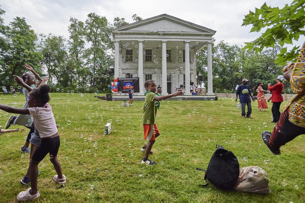 A child plays with bubbles on a lawn in front of a two-story, white farmhouse with five columns on its front facade.