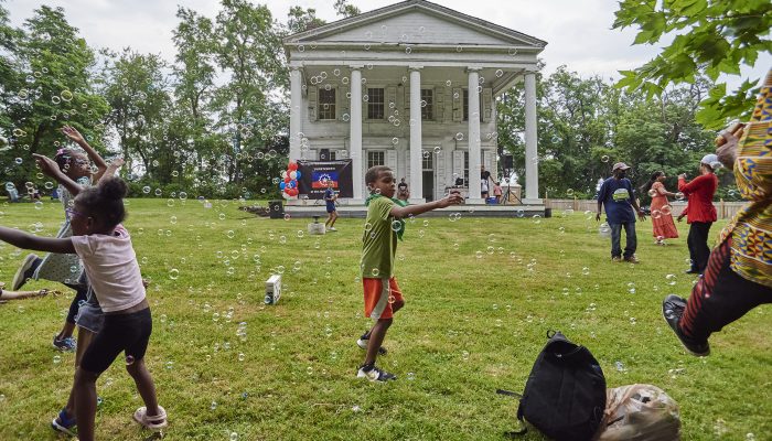 A child plays with bubbles on a lawn in front of a two-story, white farmhouse with five columns on its front facade.