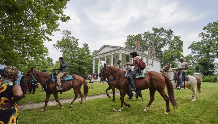 Three horses and their riders cross the grassy lawn in front of a large, white, two-story farmhouse with columns on its front facade.