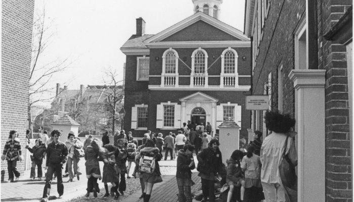 A group of young students mill about in the courtyard in front of Carpenters’ Hall and sit and stand on the stoops of the nearby recreated buildings.