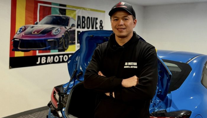 JB Motor Works’ co-founder, John Lin, stands in front of a blue car and a banner at his business