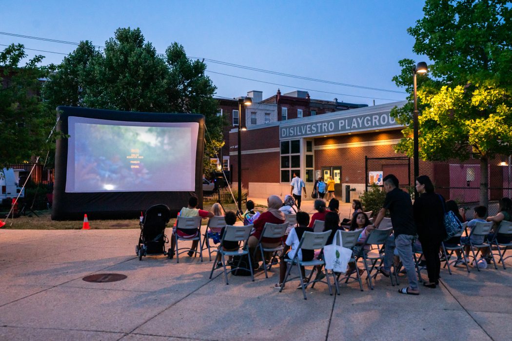 A group of neighbors seated in chairs enjoy a summer movie screening.