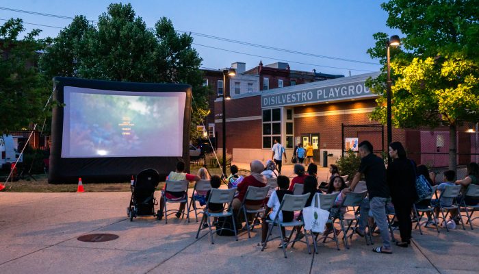 A group of neighbors seated in chairs enjoy a summer movie screening.