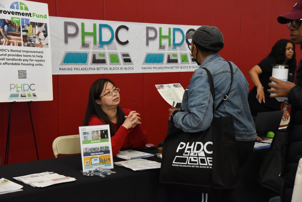 The 2023 PHDC Housing Fair. A woman sits at a table branded with PHDC logos and materials, talking to a woman carrying an informational flyer and PHDC tote bag.