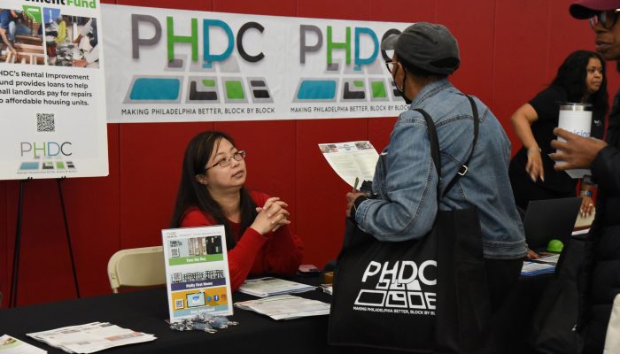 The 2023 PHDC Housing Fair. A woman sits at a table branded with PHDC logos and materials, talking to a woman carrying an informational flyer and PHDC tote bag.