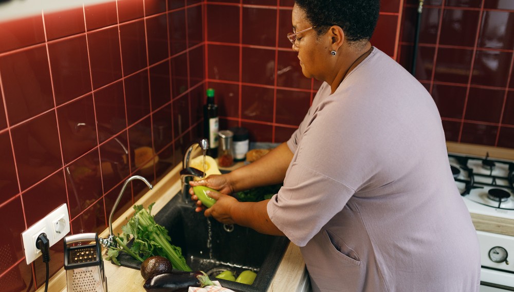 An elderly woman washes vegetables in a kitchen sink surrounded by red tiles, wearing a purple blouse and reading glasses