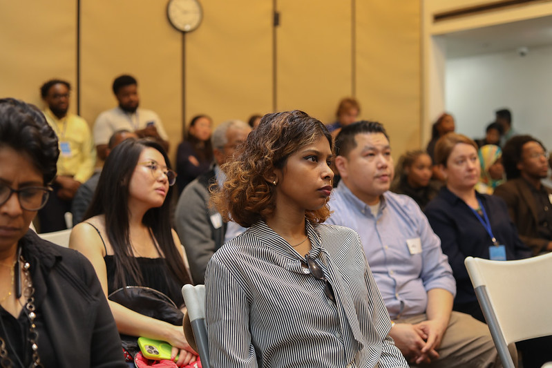 Audience members sit closely together in a room, listening to a presentation.