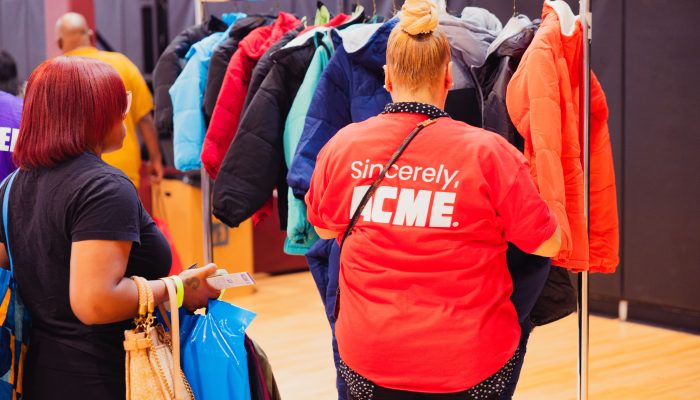 Event goers picking out winter coats from a clothing rack, while wearing red shirts that say "Sincerely, Acme" on the back.