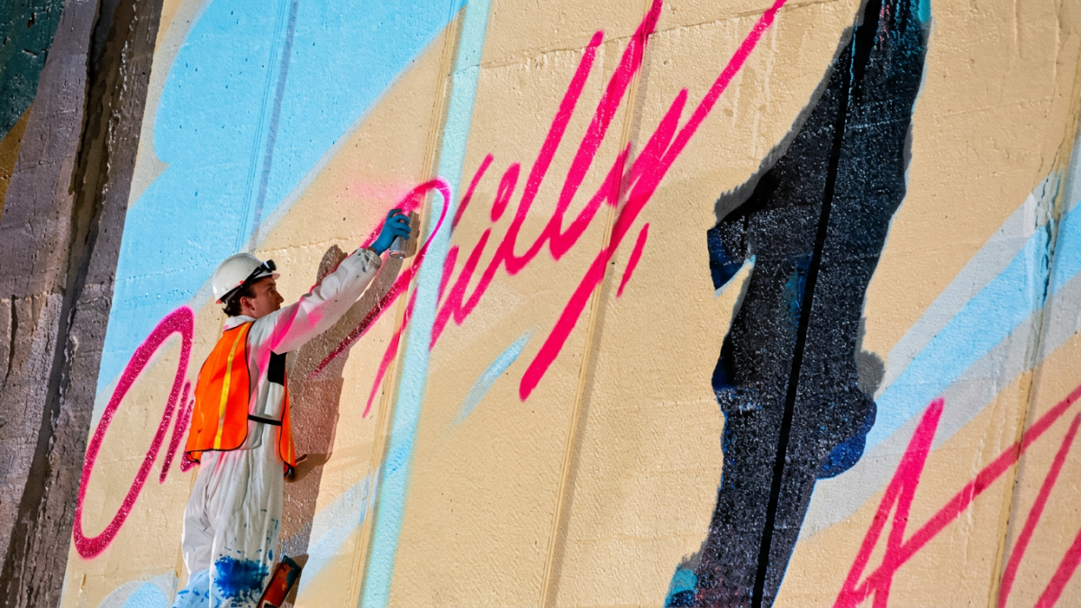 A mural artist wearing a hard hat and orange safety vest spray-paints bright pink lettering, spelling One Philly, onto a colorful Gateways Project mural in Philadelphia.