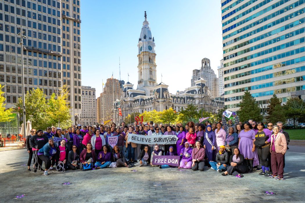 City of Philadelphia employees and community members wearing purple gather in LOVE Park with supportive banners saying "Believe Survivors" and "Freedom" in support of survivors of domestic violence