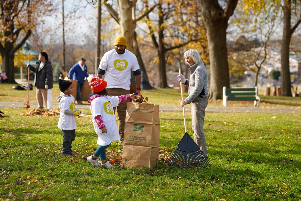 Two young children put autumn leaves into a paper lawn bag while two adults watch from the background.